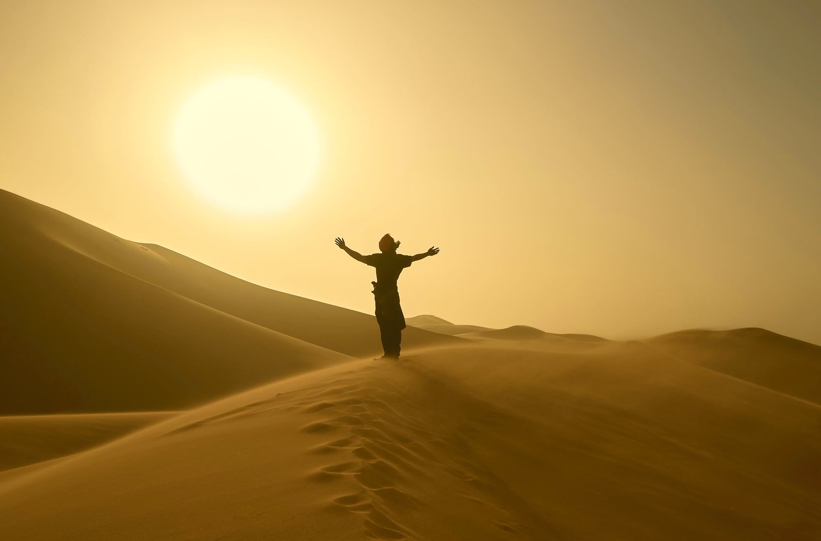 A man at the top of a golden dune, arms open to the sun, his footprints climbing behind him